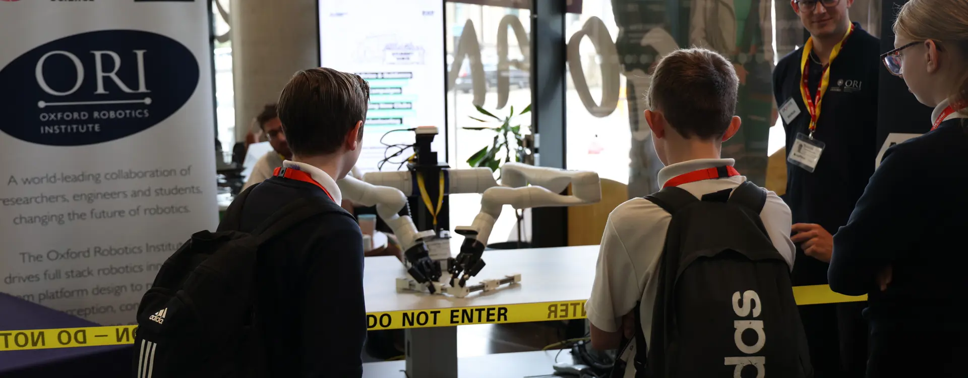 An Oxford Robotics Institute researcher stands next to Frank the robot at a demonstration booth, engaging students at the Oxplore Festival in Bradford. A branded banner and STEM posters are visible in the background.