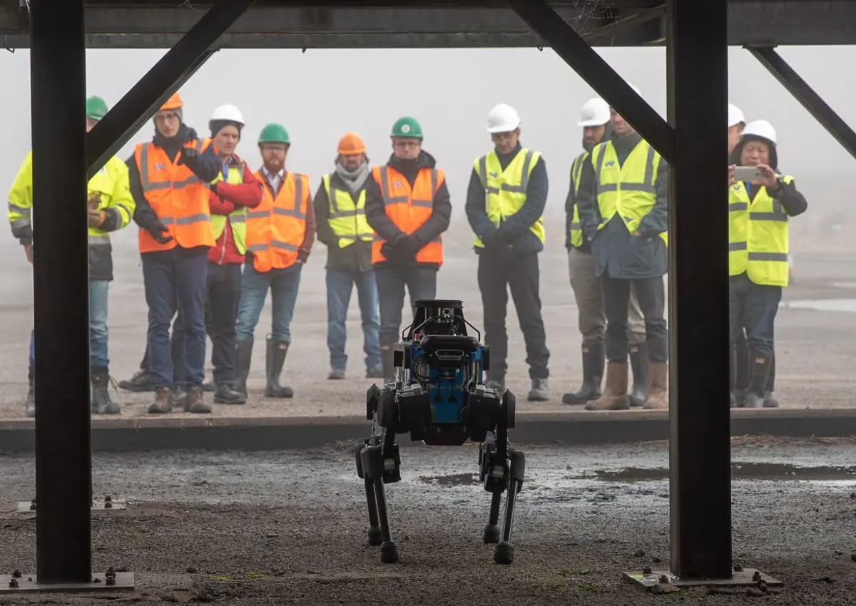 ANYmal B standing in front at industrial site, group in hivis jackets behind.